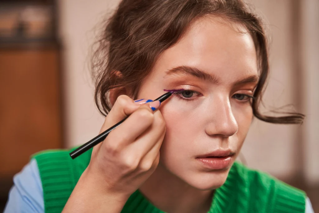 Nahaufnahme: Person trägt mit einem Stift lila Eyeliner am oberen Lid auf.