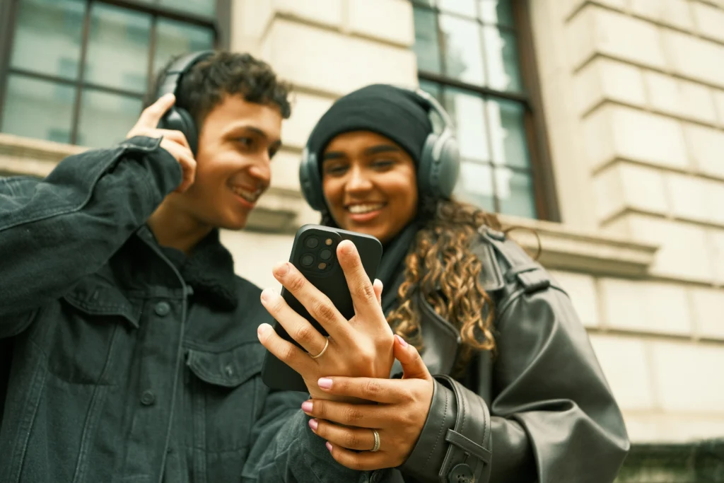 Zwei junge Menschen mit Kopfhörern schauen gemeinsam auf ein Smartphone und lächeln.