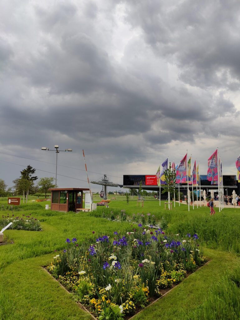 BUGA Mannheim 2023: Blumenbeet und grüne Wiesen am Gelände mit Seilbahn und bunten Fahnen vor dunklen Wolken – Stimmung und Highlights der Bundesgartenschau.
