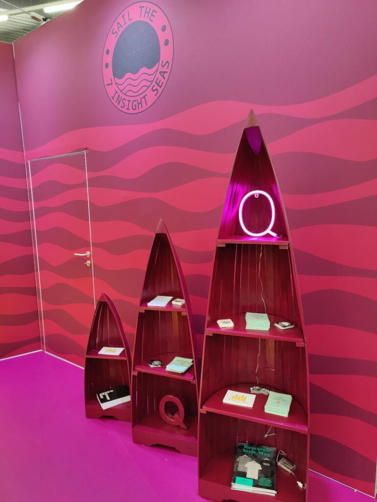 Q-Messestand auf der succeet22: pinke Wand „Sail the Insight Seas“ mit dekorativen Holzboot-Regalen und Giveaways – Standkonzept für Trendforschung und Consumer Insights.