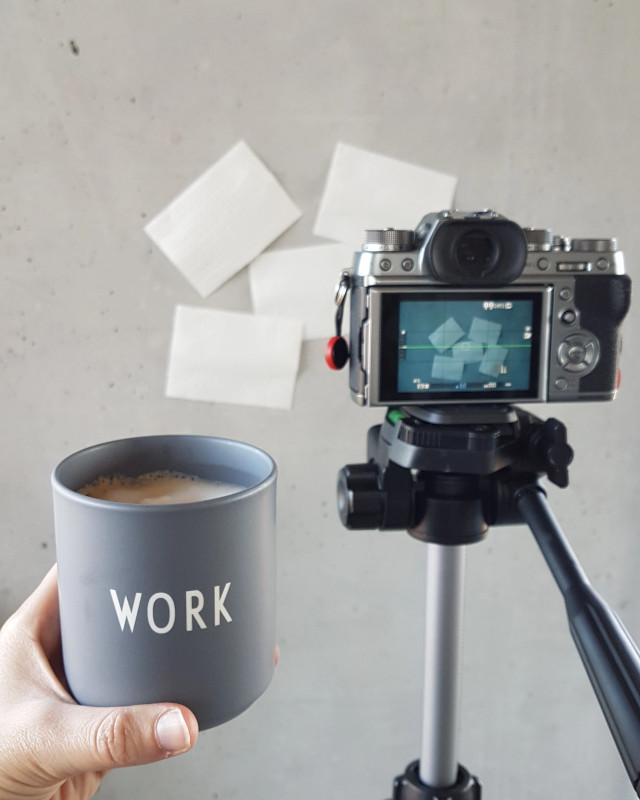 Hand hält Tasse mit Aufschrift „WORK“ neben Kamera auf Stativ vor Notizzetteln – Symbolbild für Content-Produktion, Social Media Monitoring und Q News aus der Marktforschung.